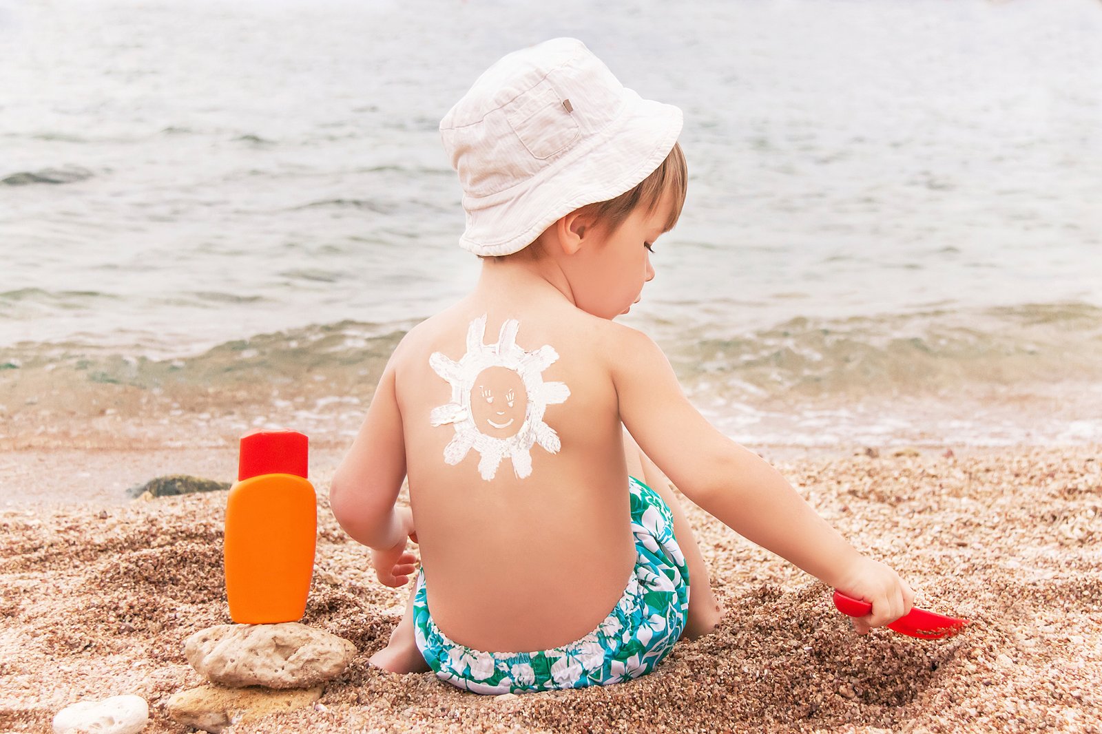 The sun drawing sunscreen (suntan lotion) on baby (boy) back. Caucasian child is sitting with plastic container of sunscreen and toy on sunny beach. Close up, outdoor (Sharm El Sheikh, Egypt).